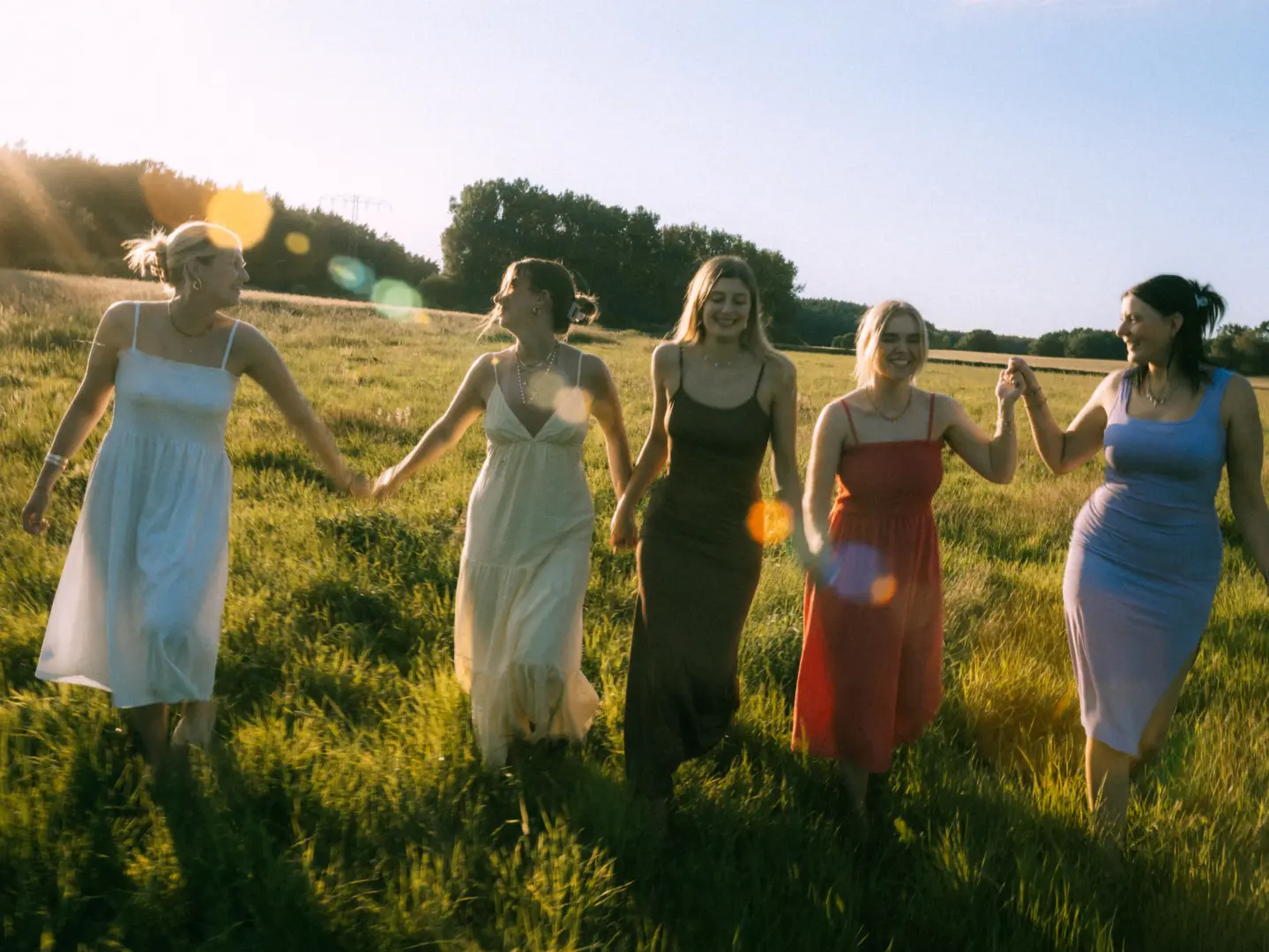 Fünf junge Frauen in sommerlichen Kleidern spazieren lachend und Händchen haltend durch eine goldene Wiese im warmen Licht der Abendsonne. Die Szene strahlt Lebensfreude und Verbundenheit aus. Ein professioneller Fotograf für Pärchen in Schwerin hält solche besonderen Momente fest.