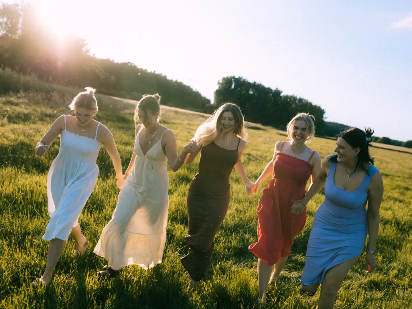 Fünf junge Frauen in sommerlichen Kleidern laufen lachend und Händchen haltend durch eine sonnendurchflutete Wiese bei Sonnenuntergang. Die Szene strahlt Lebensfreude und Verbundenheit aus. Ein stimmungsvolles Gruppenfoto für einen Fotografen für Pärchen in Schwerin.