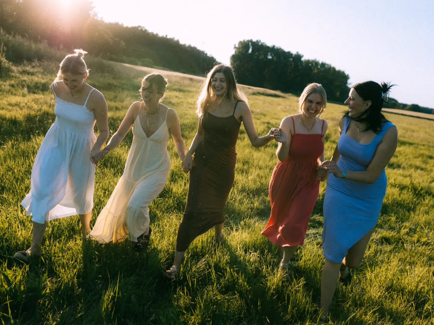Fünf junge Frauen in sommerlichen Kleidern laufen lachend und Händchen haltend durch ein sonnendurchflutetes Feld. Die warme Abendsonne sorgt für eine fröhliche Atmosphäre bei diesem Shooting, das von einem Fotograf für Pärchen in Schwerin festgehalten wurde.