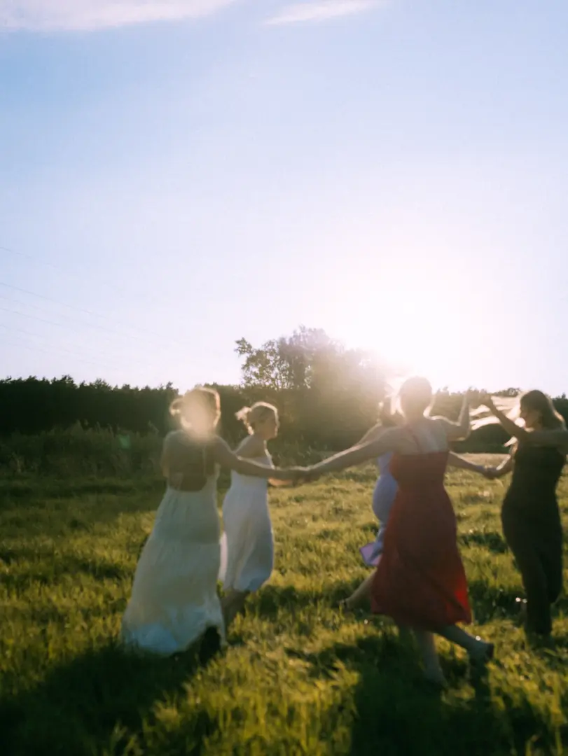 Eine Gruppe von Frauen in eleganten Sommerkleidern hält sich bei Sonnenuntergang an den Händen und läuft lachend über eine goldene Wiese. Die warme Lichtstimmung unterstreicht den fröhlichen Moment. Hochzeitsfotograf in Schwerin und MV.
