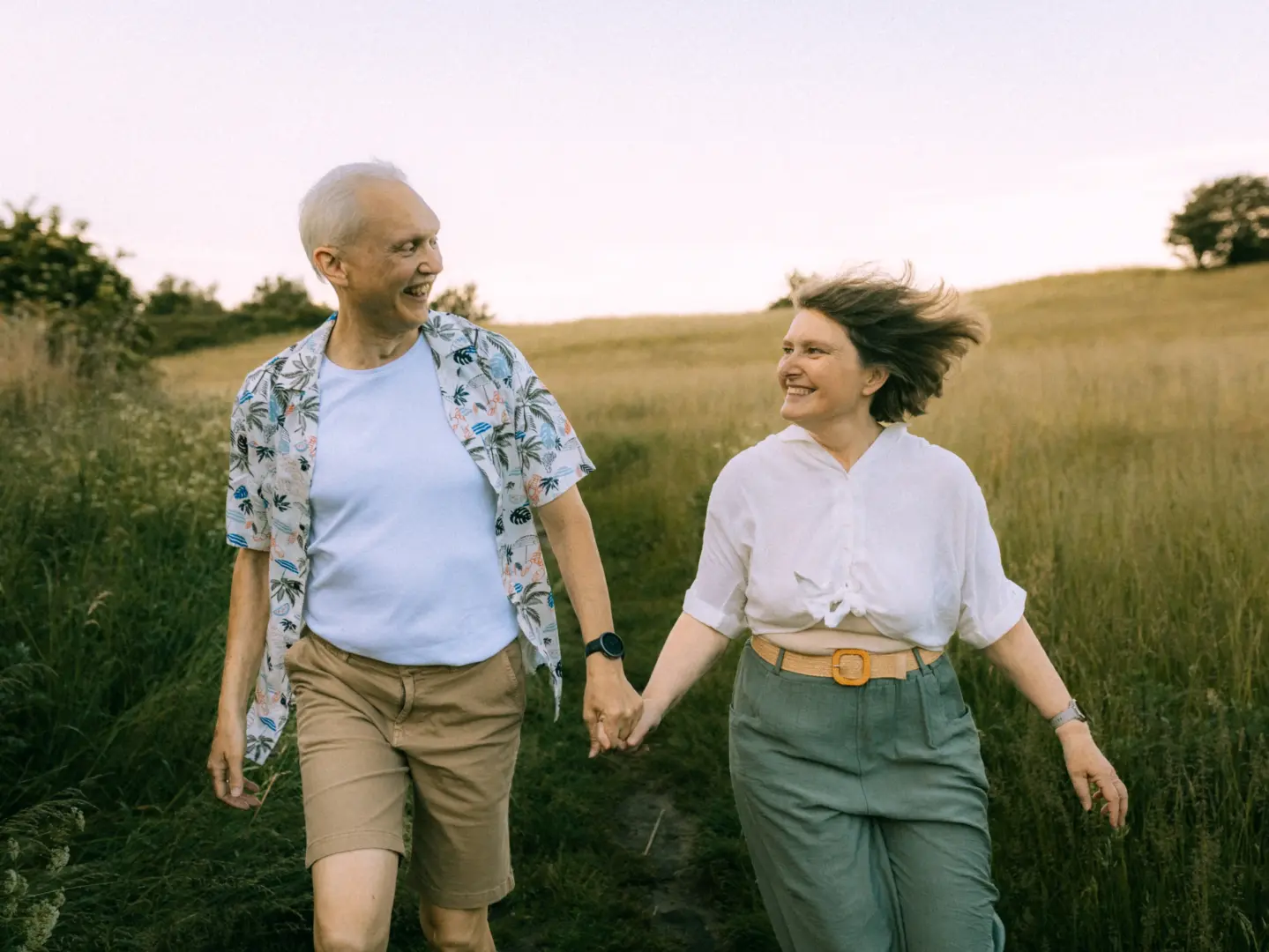 Ein glückliches, älteres Paar hält Händchen bei einem entspannten Spaziergang durch eine sommerliche Wiese bei Sonnenuntergang. Die beiden lachen sich an, während der Wind sanft durch das Haar der Frau weht. Authentische Pärchenfotos Schwerin in natürlicher Umgebung.