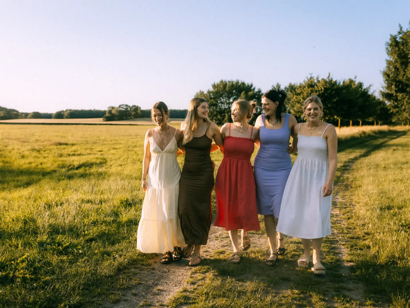 Fünf junge Frauen in sommerlichen Kleidern laufen lachend und Arm in Arm über einen Feldweg bei Sonnenuntergang. Die warme Lichtstimmung unterstreicht die freundschaftliche Atmosphäre in der Natur. Fotograf für Familien in Schwerin.