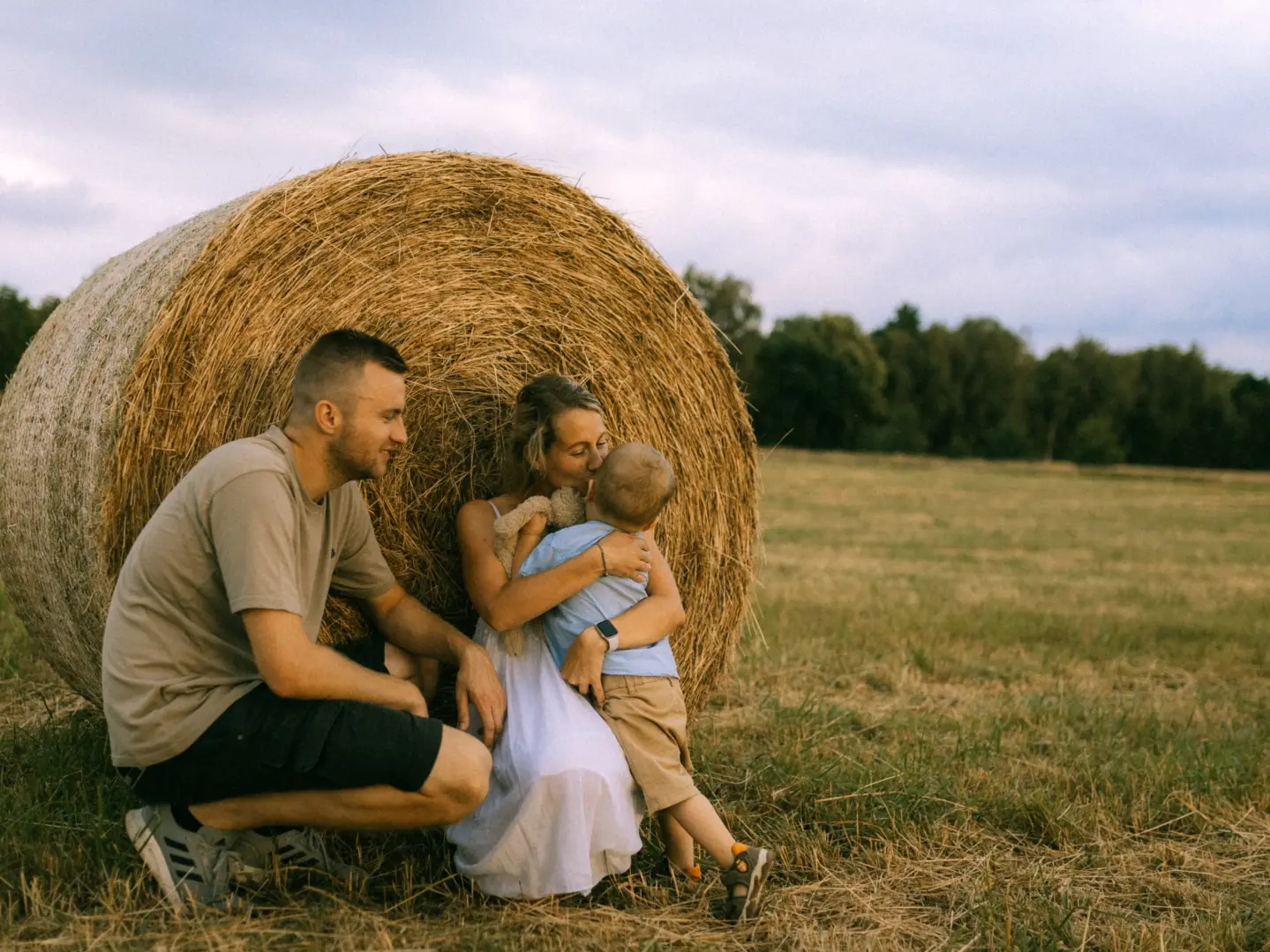 Eine junge Familie genießt einen Moment der Verbundenheit vor einem großen Heuballen auf einem abgeernteten Feld. Der Vater kniet liebevoll neben der Mutter, die ihren kleinen Sohn in den Armen hält. Ein authentisches Familienfoto für den Fotograf für Familien in Schwerin.