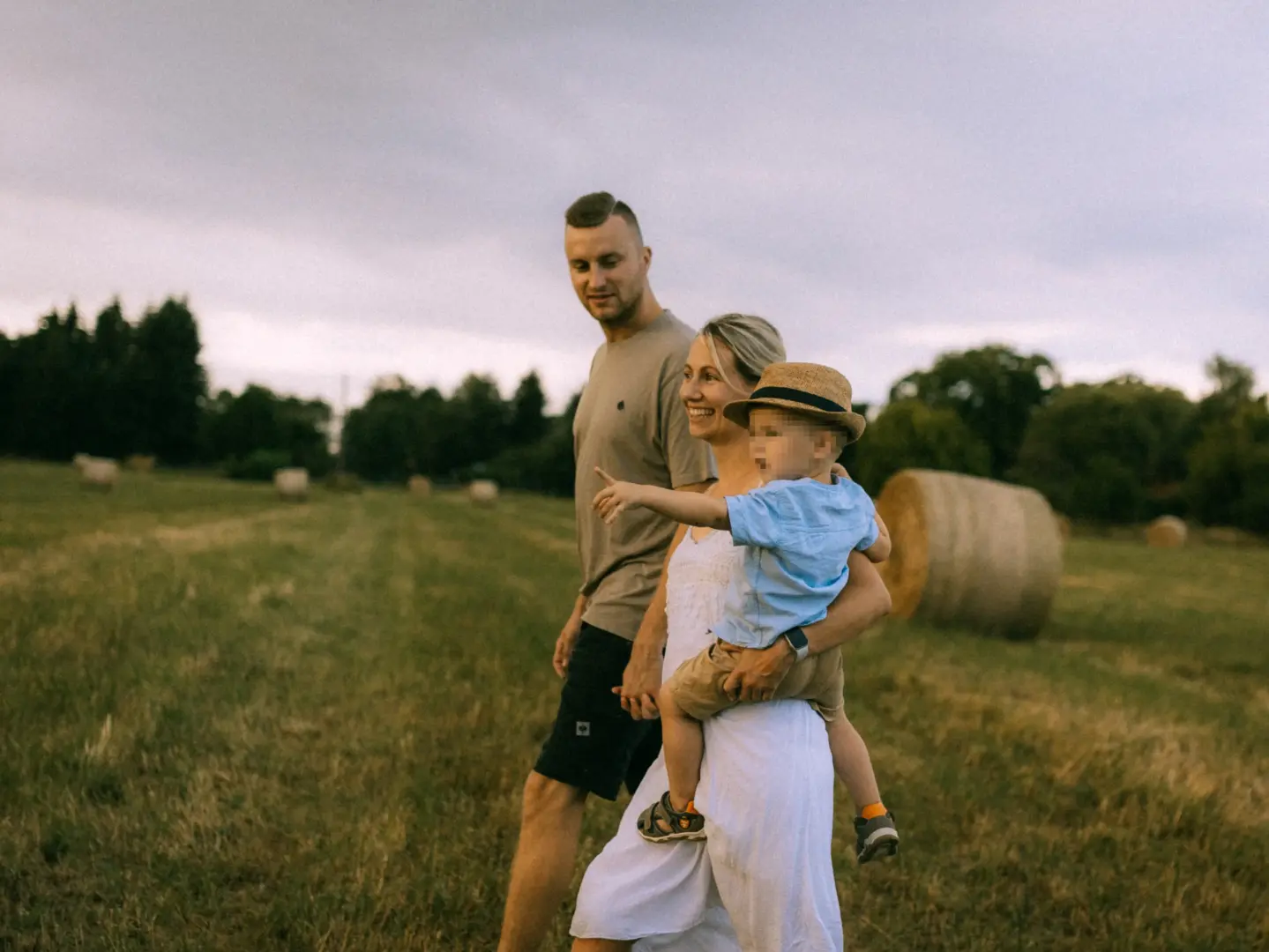 Ein glückliches Paar spaziert bei Sonnenuntergang mit ihrem kleinen Sohn auf dem Arm über ein Feld mit Heuballen. Der Junge trägt einen Strohhut und zeigt in die Ferne. Authentische Familienfotos in Schwerin, die eine warme und liebevolle Stimmung einfangen.