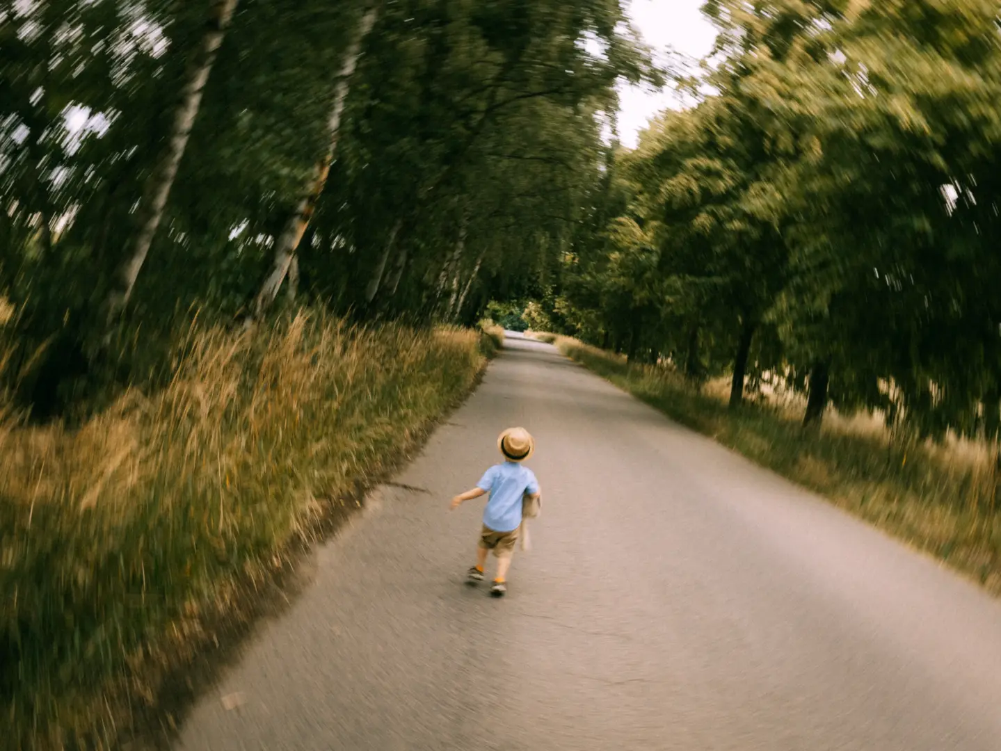 Ein kleines Kind mit Strohhut und blauem Hemd läuft fröhlich einen von Bäumen gesäumten Weg entlang, wobei der Hintergrund einen dynamischen Bewegungsunschärfe-Effekt aufweist. Familienfotograf in Schwerin.