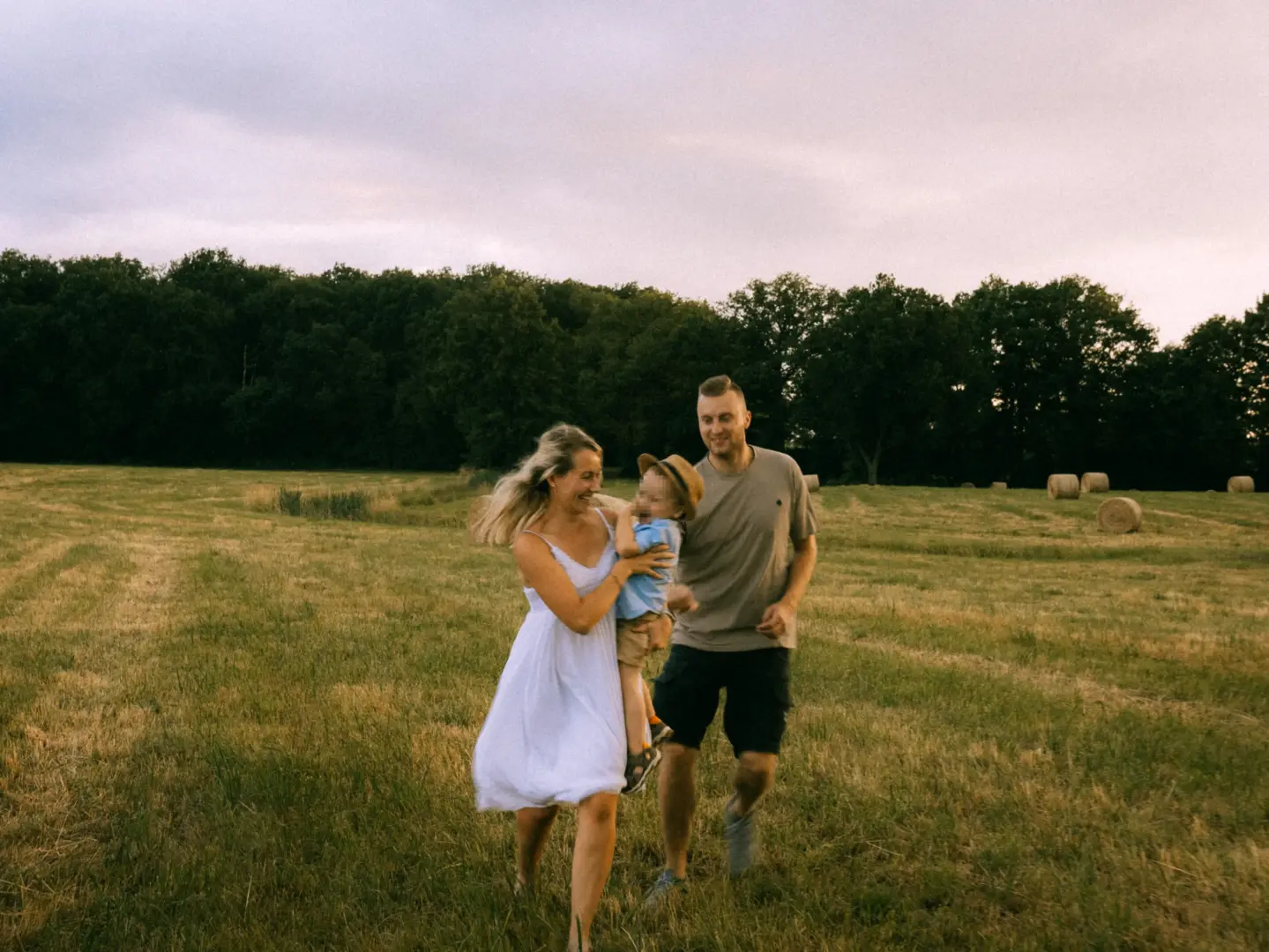Eine junge Familie läuft lachend über ein goldenes Stoppelfeld bei Sonnenuntergang. Die Mutter trägt ein weißes Sommerkleid und hält ihren kleinen Sohn auf dem Arm, während der Vater neben ihnen hergeht. Im Hintergrund sind ein Waldrand und Strohballen zu sehen. Familienfotograf in Schwerin.