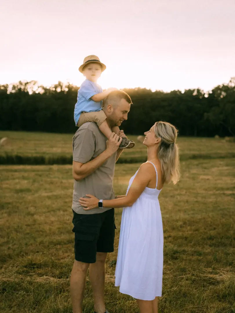 Eine junge Familie genießt einen sonnigen Moment auf einem Feld bei Sonnenuntergang. Der Vater trägt seinen Sohn auf den Schultern, während die Mutter ihn liebevoll ansieht. Ein herzliches Porträt von einem professionellen Familienfotograf in Schwerin.
