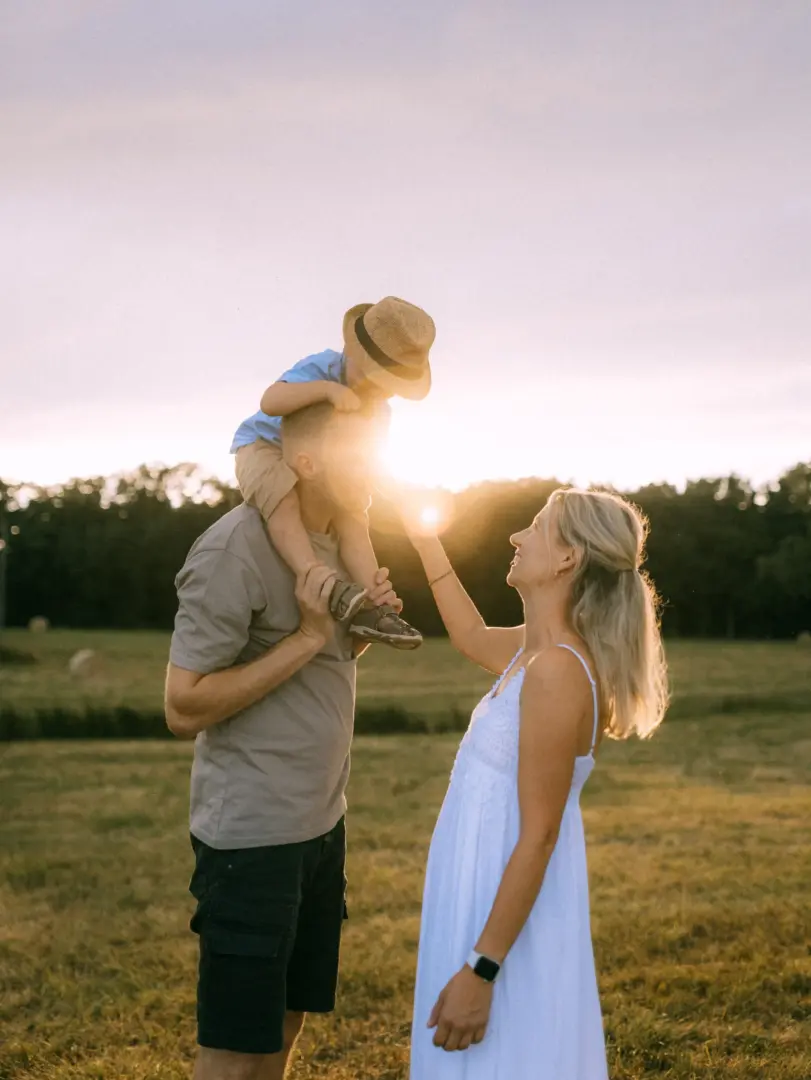 Ein glückliches Paar mit ihrem kleinen Sohn auf den Schultern bei einem sonnigen Spaziergang auf einem Feld. Die warme Abendsonne sorgt für eine stimmungsvolle Atmosphäre. Professionelle Familienfotos in Schwerin, die natürliche Momente voller Liebe festhalten.