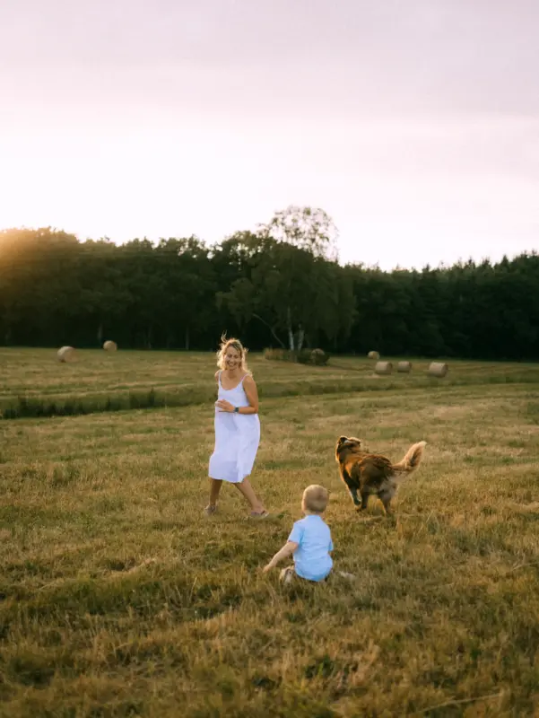 Eine lächelnde Mutter in einem weißen Sommerkleid spielt bei goldenem Sonnenuntergang mit ihrem kleinen Sohn und ihrem Hund auf einer weitläufigen Wiese mit Heuballen. Ein herzlicher Moment für Familienfotos in Schwerin.