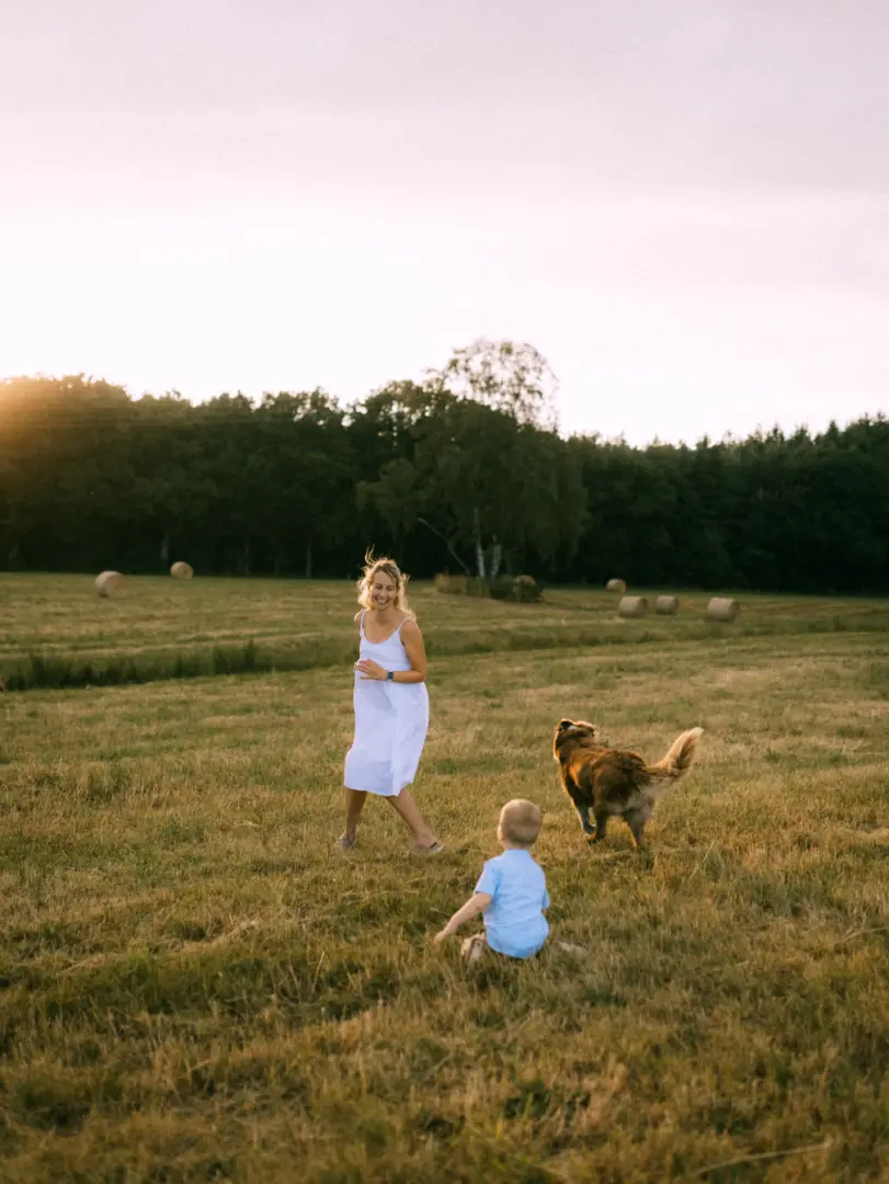 Eine lächelnde Mutter in einem weißen Sommerkleid spielt bei goldenem Sonnenuntergang mit ihrem kleinen Sohn und ihrem Hund auf einer weitläufigen Wiese mit Heuballen. Ein herzlicher Moment für Familienfotos in Schwerin.