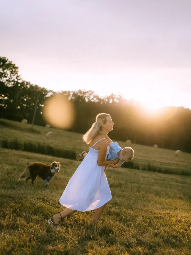 Eine Mutter in einem weißen Sommerkleid spielt glücklich mit ihrem kleinen Sohn auf einer sonnenverwöhnten Wiese bei Sonnenuntergang, während ihr Hund im Hintergrund läuft. Authentische Familienfotos in Schwerin, die eine liebevolle Bindung in der Natur einfangen.