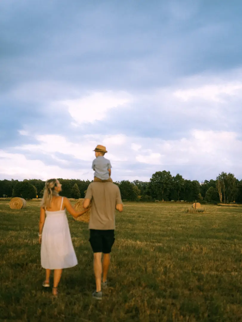 Eine junge Familie bei einem Spaziergang über ein goldenes Stoppelfeld im warmen Abendlicht. Ein Vater trägt seinen kleinen Sohn auf den Schultern, während die Mutter neben ihnen hergeht. Familienfotograf in Schwerin, der natürliche Momente im Freien einfängt.