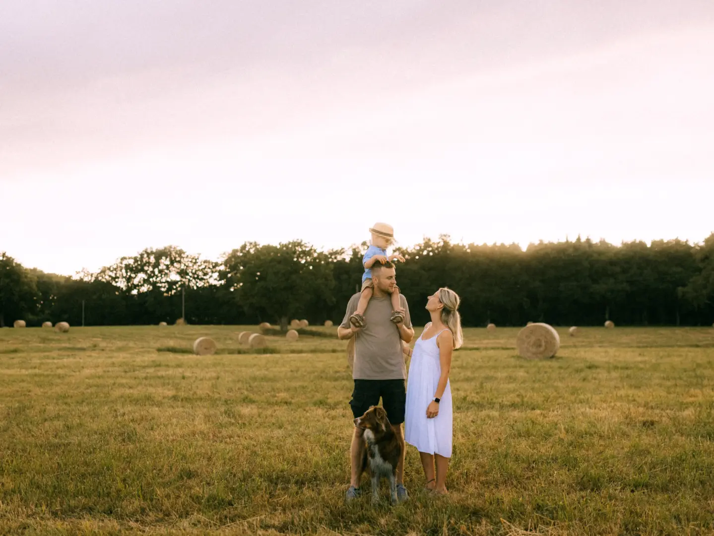 Eine junge Familie genießt einen sonnigen Abend auf einem Feld mit Heuballen. Der Vater trägt seinen kleinen Sohn auf den Schultern, während die Mutter neben ihm steht und ihn anlächelt. Ein treuer Hund begleitet sie. Authentische Familienfotos in Schwerin bei goldenem Licht.