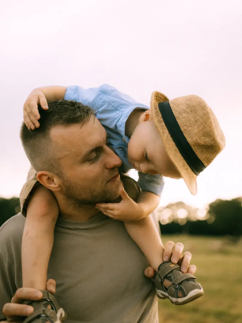 Ein liebevoller Vater trägt seinen kleinen Sohn auf den Schultern durch eine sonnige Wiese. Das Kind trägt einen Strohhut und schaut den Vater zärtlich an, während dieser lächelnd den Blick erwidert. Ein herzlicher Moment für Familienfotos in Schwerin.