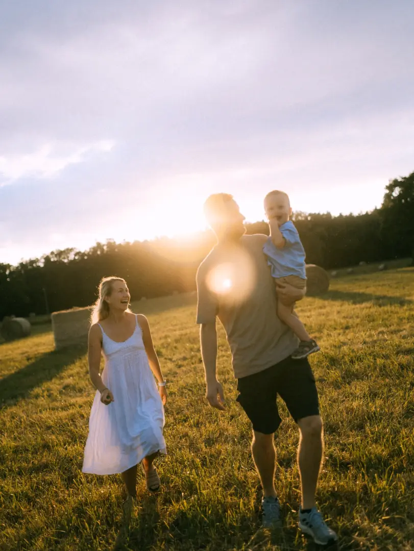 Eine junge Familie genießt einen sonnigen Moment auf einem Feld mit Heuballen. Ein Familienfotograf in Schwerin hat diesen herzlichen Augenblick eingefangen, als der Vater sein Kind auf dem Arm trägt und die Mutter glücklich neben ihnen herläuft, während die Sonne warmes Licht spendet.