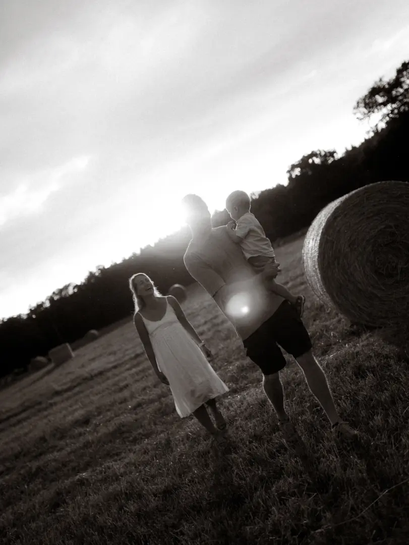Eine junge Familie genießt einen sonnigen Moment auf einem Feld mit Heuballen. Ein Vater trägt sein Kleinkind auf dem Arm, während die Mutter glücklich neben ihnen hergeht. Ein stimmungsvolles Familienfoto, aufgenommen von einem Fotograf für Familien in Schwerin.
