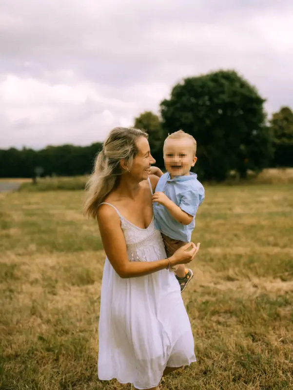 Eine strahlende Mutter im weißen Sommerkleid trägt ihren kleinen Sohn lachend auf dem Arm. Sie spazieren gemeinsam über eine herbstliche Wiese unter bewölktem Himmel. Authentische Familienfotos in Schwerin, die eine herzliche Verbindung und unbeschwerte Lebensfreude einfangen.