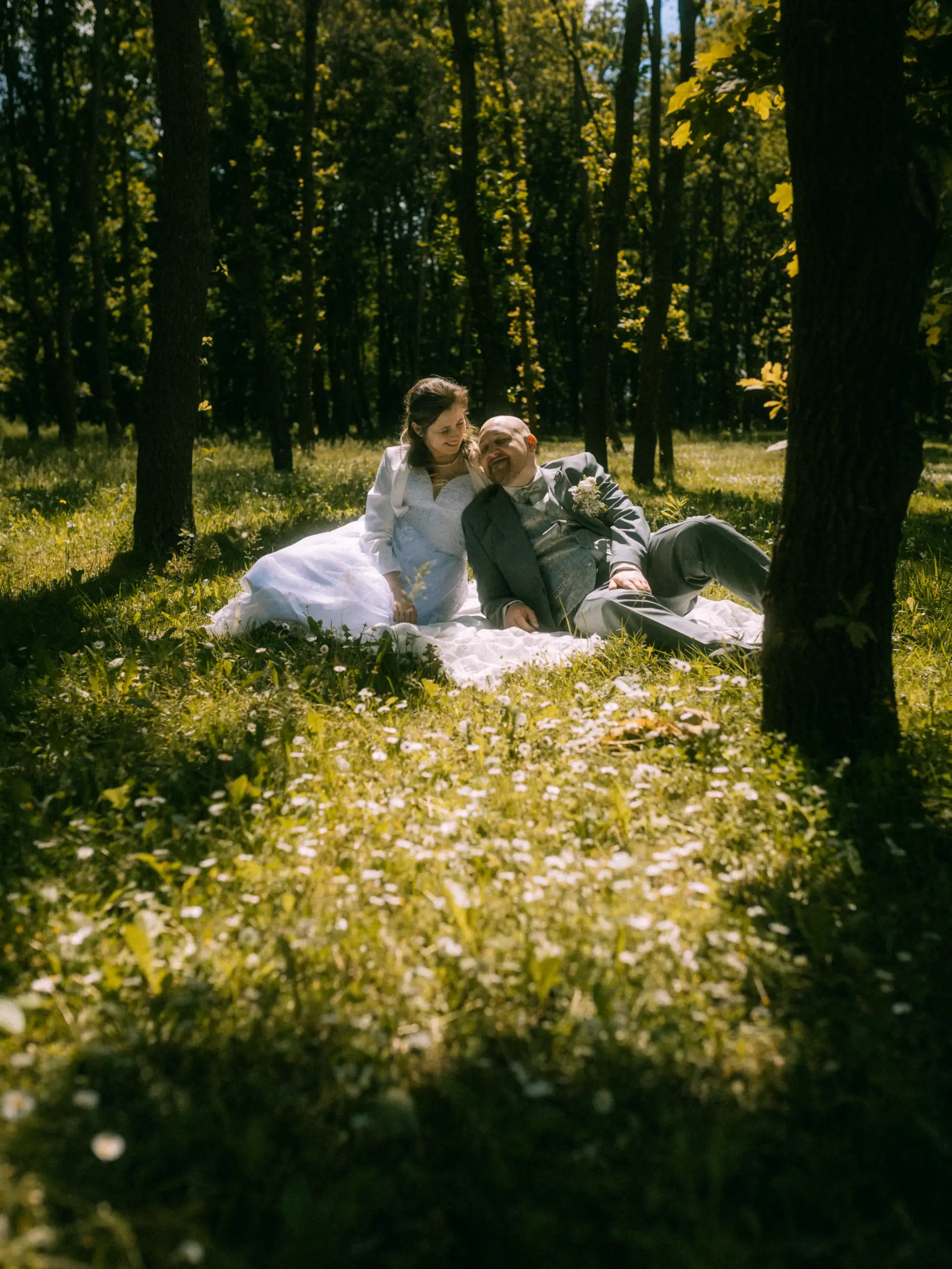 Ein glückliches Brautpaar sitzt entspannt auf einer Picknickdecke inmitten einer sonnendurchfluteten Waldlichtung voller Gänseblümchen. Die Braut trägt ein weißes Kleid, der Bräutigam einen grauen Anzug. Eine romantische Szene, eingefangen von einem Hochzeitsfotograf in Schwerin und MV.