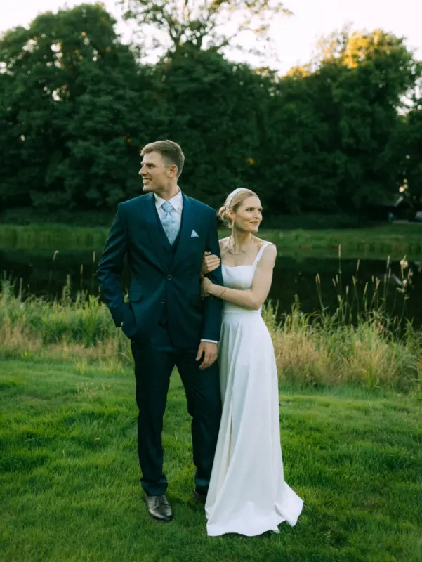 Ein Brautpaar posiert für Pärchenfotos in Schwerin: Der Bräutigam trägt einen eleganten dunkelblauen Anzug, während die Braut in einem schlichten weißen Hochzeitskleid mit Haarband an seinem Arm lehnt. Beide blicken verträumt in die Ferne vor einer idyllischen See- und Waldkulisse.