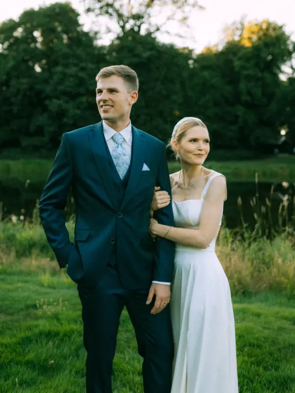 Ein Brautpaar posiert glücklich im Freien an einem Seeufer. Der Bräutigam trägt einen eleganten dunkelblauen Anzug, die Braut ein schlichtes weißes Kleid mit Kopfband. Das stimmungsvolle Licht der Abendsonne unterstreicht diese romantische Lovestory als Fotograf für Pärchen in Schwerin.