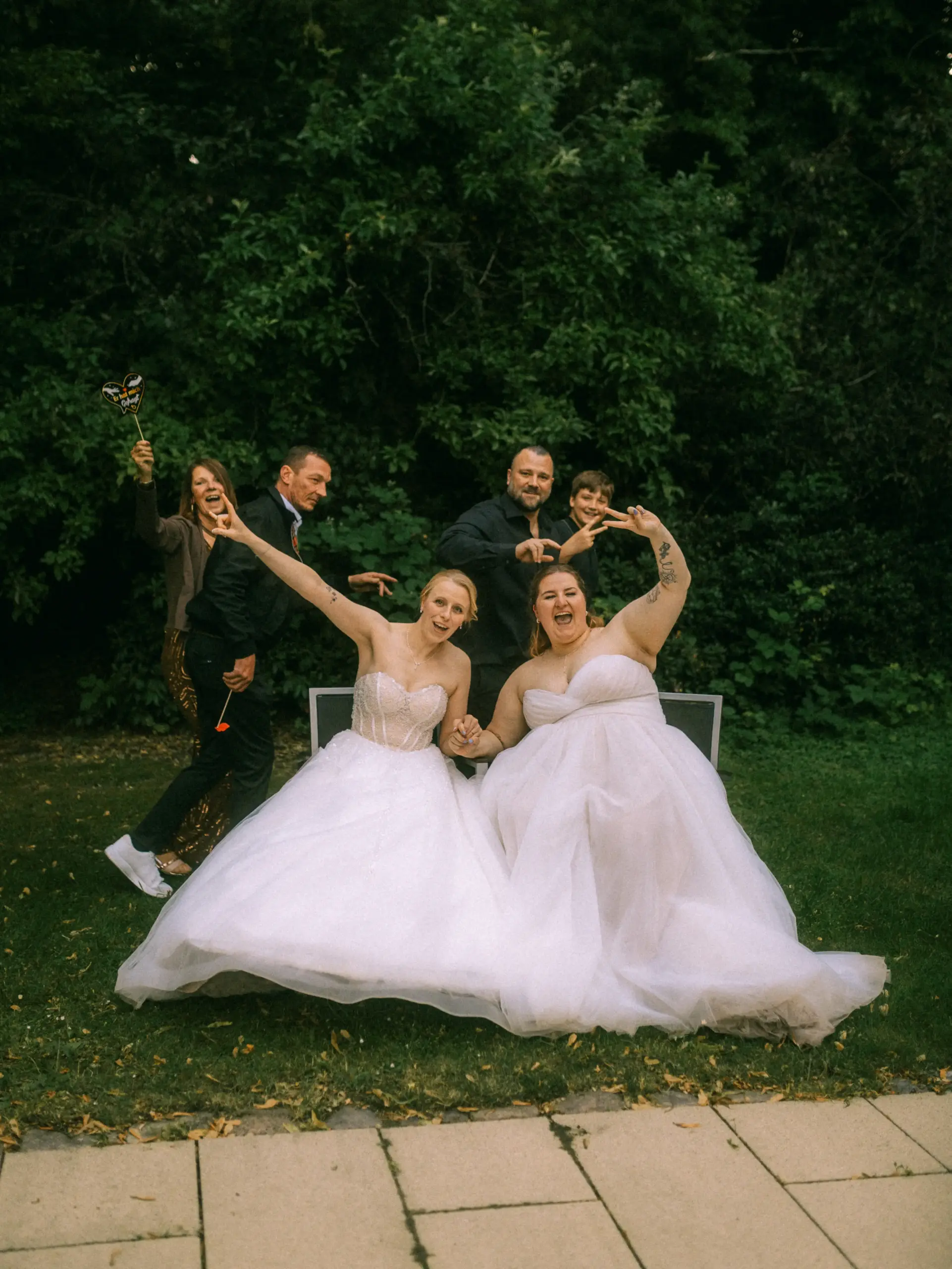 Zwei strahlende Bräute in weißen Hochzeitskleidern sitzen auf einer Bank und feiern ausgelassen mit ihren Gästen im Grünen. Ein Hochzeitsfotograf in Schwerin und MV hat diesen lebendigen Moment voller Freude und Spaß perfekt eingefangen.