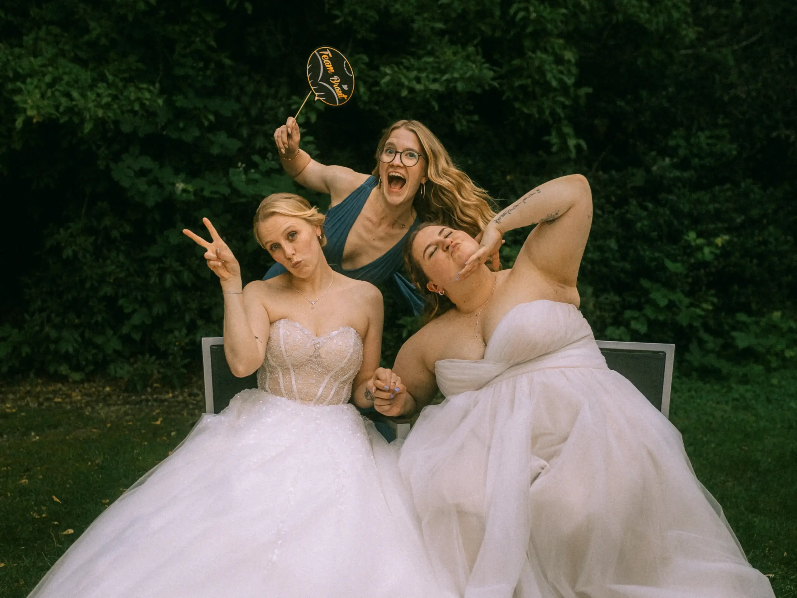 Zwei Bräute in eleganten weißen Hochzeitskleidern machen gemeinsam mit einer Freundin, die ein „Team Bräute“-Schild hält, lustige Grimassen für ein Foto. Die ausgelassene Stimmung fängt die Arbeit als Hochzeitsfotograf in Schwerin und MV perfekt ein. Zwei Bräute in eleganten weißen Hochzeitskleidern machen gemeinsam mit einer Freundin, die ein „Team Bräute“-Schild hält, lustige Grimassen für ein Foto. Die ausgelassene Stimmung fängt die Arbeit als Hochzeitsfotograf in Schwerin und MV perfekt ein.