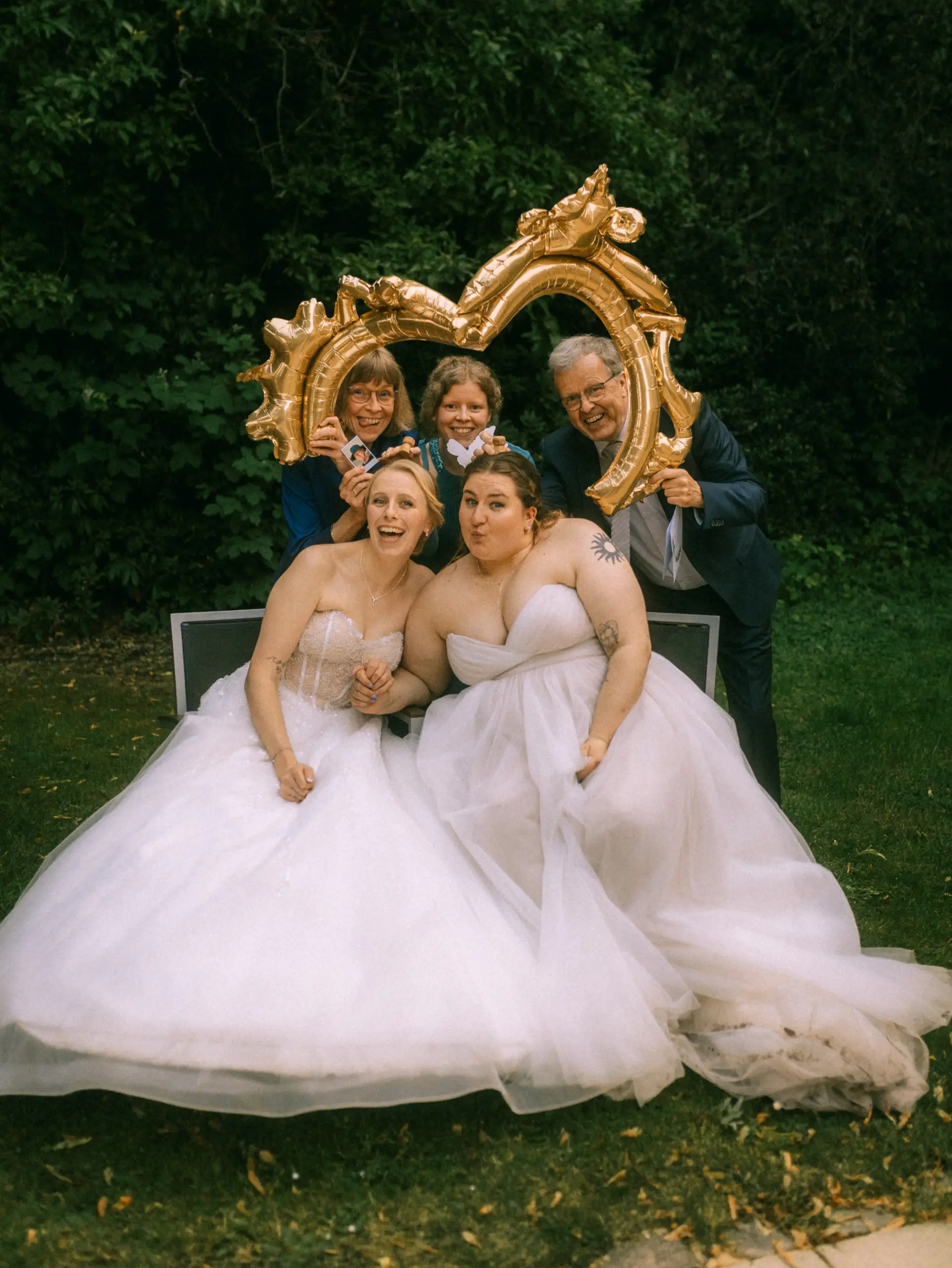 Zwei glückliche Bräute in weißen Brautkleidern sitzen auf einer Bank, umrahmt von ihren Eltern, die einen goldenen herzförmigen Luftballon halten. Ein fröhliches Hochzeitsfoto, das als Hochzeitsfotograf in Schwerin und MV festgehalten wurde.