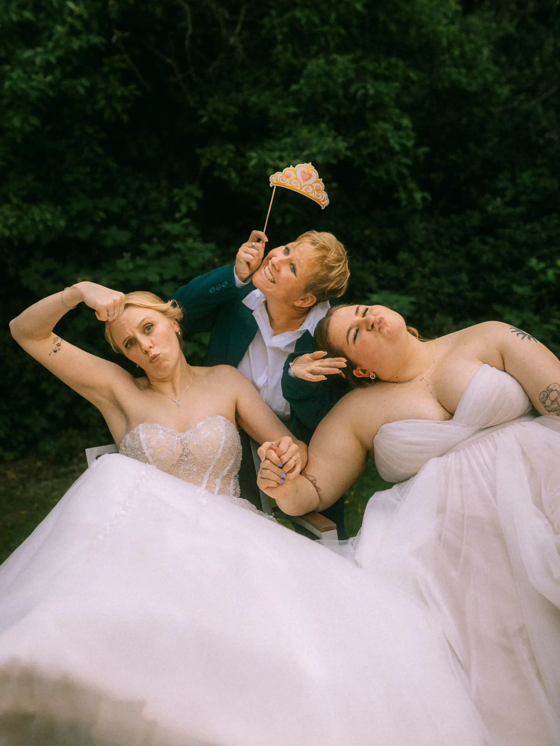 Zwei Bräute in weißen Brautkleidern sitzen zusammen mit einer Person in einem grünen Anzug, die eine Kronen-Requisite über ihre Köpfe hält. Alle drei machen lustige Gesichter in einer entspannten Outdoor-Umgebung. Stimmungsvolle Aufnahme von einem Hochzeitsfotograf in Schwerin und MV. Zwei Bräute in weißen Brautkleidern sitzen zusammen mit einer Person in einem grünen Anzug, die eine Kronen-Requisite über ihre Köpfe hält. Alle drei machen lustige Gesichter in einer entspannten Outdoor-Umgebung. Stimmungsvolle Aufnahme von einem Hochzeitsfotograf in Schwerin und MV.