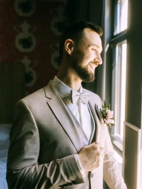 Ein glücklicher Bräutigam mit Bart in einem eleganten beigen Hochzeitsanzug mit Fliege und Blumenanstecker blickt lächelnd aus einem Fenster im Schloss Basthorst vor der Hochzeit, eingefangen von einem Hochzeitsfotograf in Crivitz.
