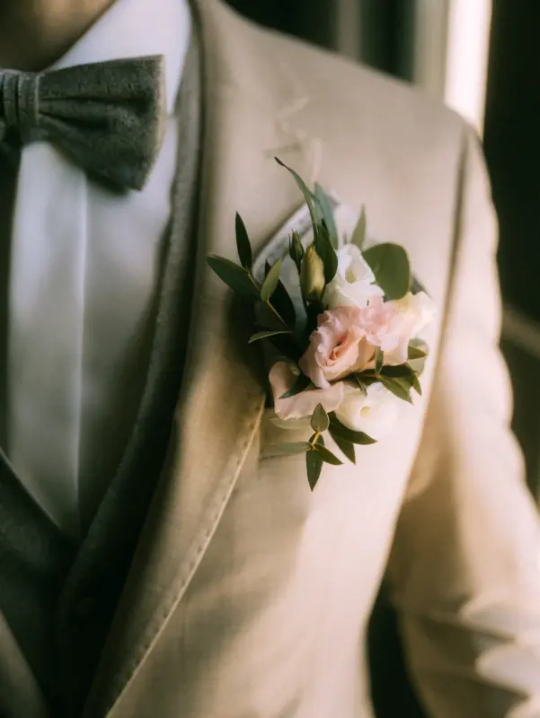 Ein Bräutigam in einem beigen Anzug mit einer eleganten Weste und einer gemusterten Fliege trägt ein florales Ansteckblümchen aus zarten rosa und weißen Rosen sowie Eukalyptusblättern. Detailaufnahme für einen Hochzeitsfotograf in Schwerin und MV.
