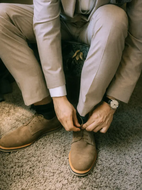 Ein Bräutigam bereitet sich auf seine Hochzeit vor und bindet sich die beigen Wildlederschuhe, während er einen eleganten beigen Anzug trägt. Der Moment wird von einem Hochzeitsfotograf in Schwerin und MV festgehalten, mit Fokus auf die Details wie die Armbanduhr und den Teppichboden.