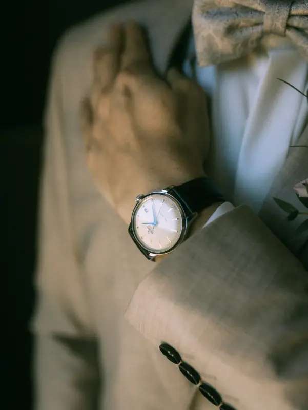 Nahaufnahme eines Bräutigams in einem beigen Anzug mit Fliege, der eine elegante Orient-Armbanduhr mit blauem Sekundenzeiger trägt. Das stilvolle Detail unterstreicht die festliche Stimmung, eingefangen von Ihrem Hochzeitsfotograf in Schwerin und MV.