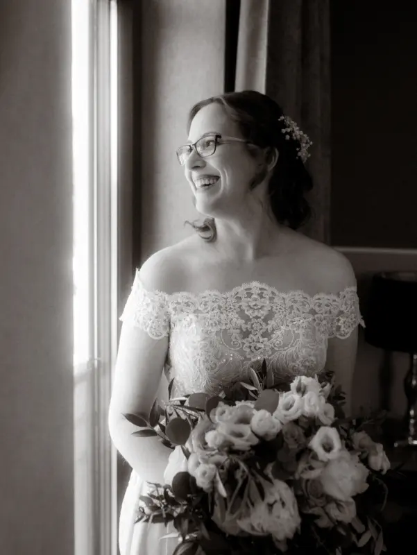 Eine strahlende Braut in einem eleganten schulterfreien Spitzenkleid hält einen Blumenstrauß in den Händen und blickt glücklich aus einem Fenster vom Schloss Basthorst. Die Aufnahme im Vintage-Stil unterstreicht die emotionale Stimmung als Hochzeitsfotograf in Crivitz.