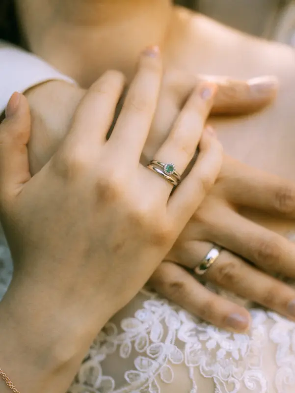 Nahaufnahme eines glücklichen Brautpaares, wobei die Hände zärtlich ineinander verschränkt sind. Die Braut trägt einen eleganten Verlobungsring mit grünem Stein und einen Ehering, während das Paar vor einem weißen Spitzenkleid posiert. Romantische Pärchenfotos Schwerin.