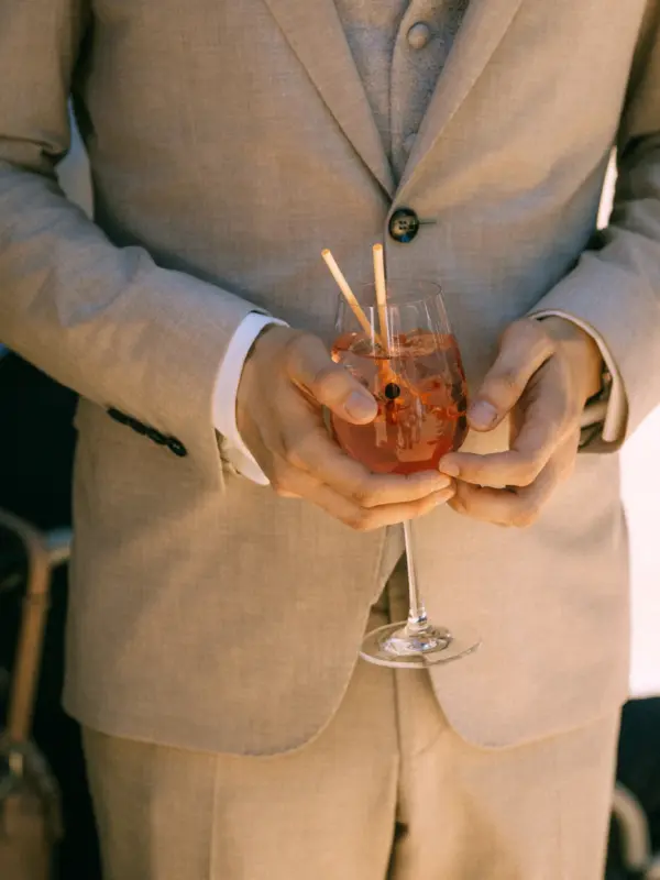 Ein eleganter Bräutigam in einem beigen Anzug hält ein Glas mit einem erfrischenden Aperol Spritz in den Händen. Die Aufnahme fängt die stilvolle Atmosphäre einer Hochzeit ein, festgehalten von einem Hochzeitsfotograf in Schwerin und MV.