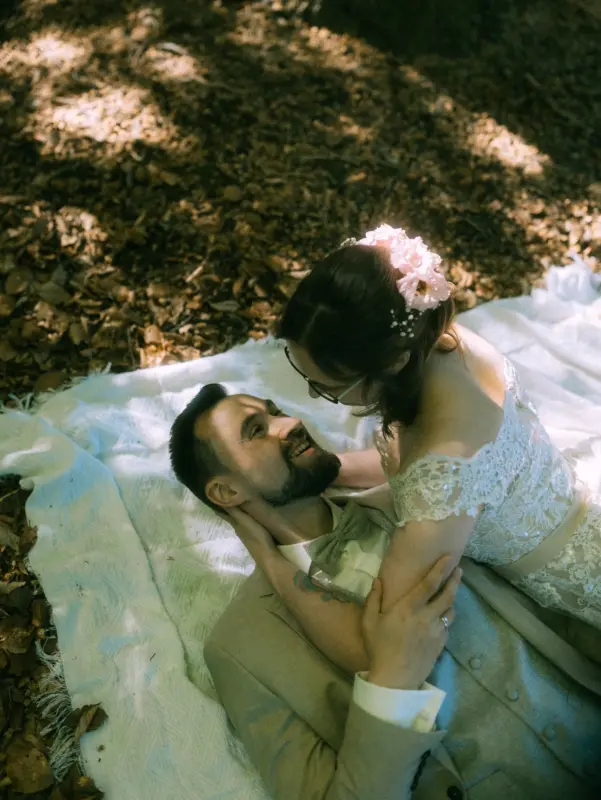 Ein verliebtes Brautpaar liegt auf einer Picknickdecke im Wald nahe Schloss Basthorst bei Crivitz. Die Braut mit Blumen im Haar hält zärtlich das Gesicht des Bräutigams im beigen Anzug. Romantische Hochzeitsaufnahme, fotografiert von Hochzeitsfotograf Jan Büchler aus Crivitz.