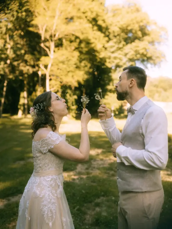 Ein Brautpaar bläst gemeinsam Pusteblumen im Garten von Schloss Basthorst bei Crivitz. Die Braut trägt ein weißes Spitzenkleid mit Blumen im Haar, der Bräutigam eine Weste mit Fliege. Dieses romantische Hochzeitsfoto wurde vom Hochzeitsfotografen Jan Büchler aus Crivitz aufgenommen.