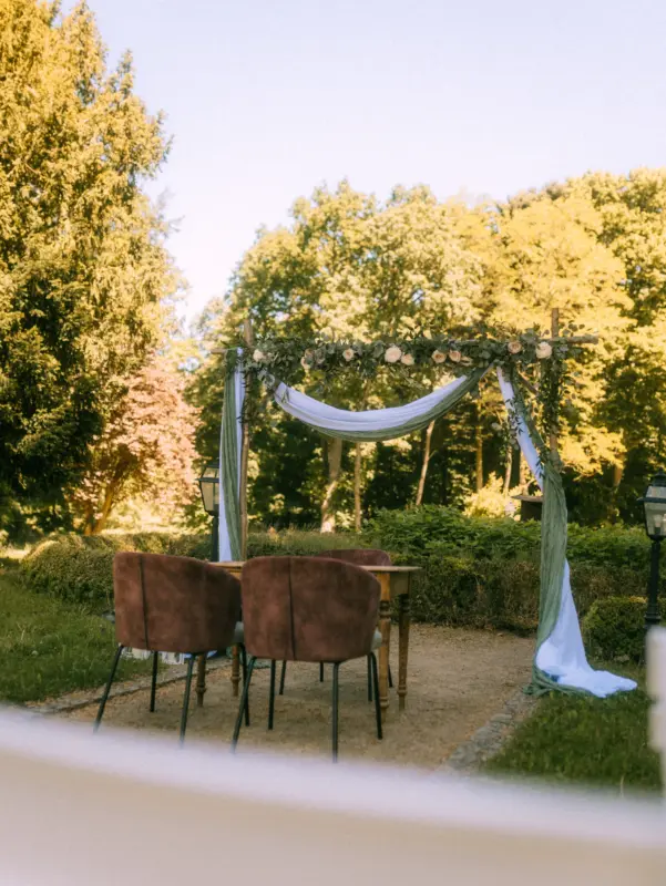 Ein eleganter Traubogen mit Blumen und fließenden Stoffen steht im Garten, davor ein Holztisch mit zwei braunen Samtstühlen für eine freie Trauung im Grünen. Eine stimmungsvolle Kulisse für Paare, eingefangen von einem Hochzeitsfotograf in Schwerin und MV.