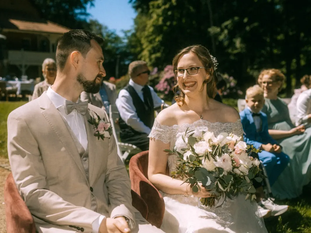 Ein glückliches Brautpaar sitzt während der Hochzeitszeremonie im Freien auf roten Sesseln. Die Braut hält einen Blumenstrauß, während sie den Bräutigam liebevoll ansieht. Im Hintergrund sitzen Gäste. Professionelle Aufnahmen vom Hochzeitsfotograf in Crivitz.