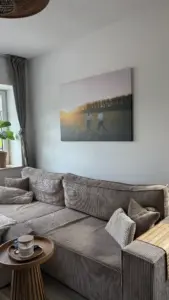 Ein großes Leinwandbild über einem beigen Cordsofa zeigt ein Paar bei Sonnenuntergang am Schloss Basthorst. Das stimmungsvolle Hochzeitsfoto wurde vom Hochzeitsfotografen Jan Büchler aus Crivitz aufgenommen und verleiht dem gemütlichen Wohnzimmer eine persönliche Note.